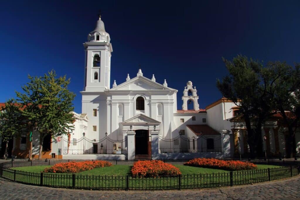 A imagem retrata a imponente fachada da Basílica de Nossa Senhora do Pilar, localizada no bairro da Recoleta, em Buenos Aires. A igreja, construída em estilo barroco colonial, apresenta uma fachada predominantemente branca, que se destaca contra o céu azul profundo.
No centro, a entrada principal é simples e elegante, com uma porta de madeira encimada por um frontão triangular. O campanário, localizado à esquerda, é alto e esguio, com detalhes ornamentais e janelas que permitem a passagem de luz. À direita da estrutura principal, há uma torre menor com um sino, complementando a simetria da construção.
À frente da basílica, o jardim bem cuidado é adornado com um canteiro de flores vibrantes.