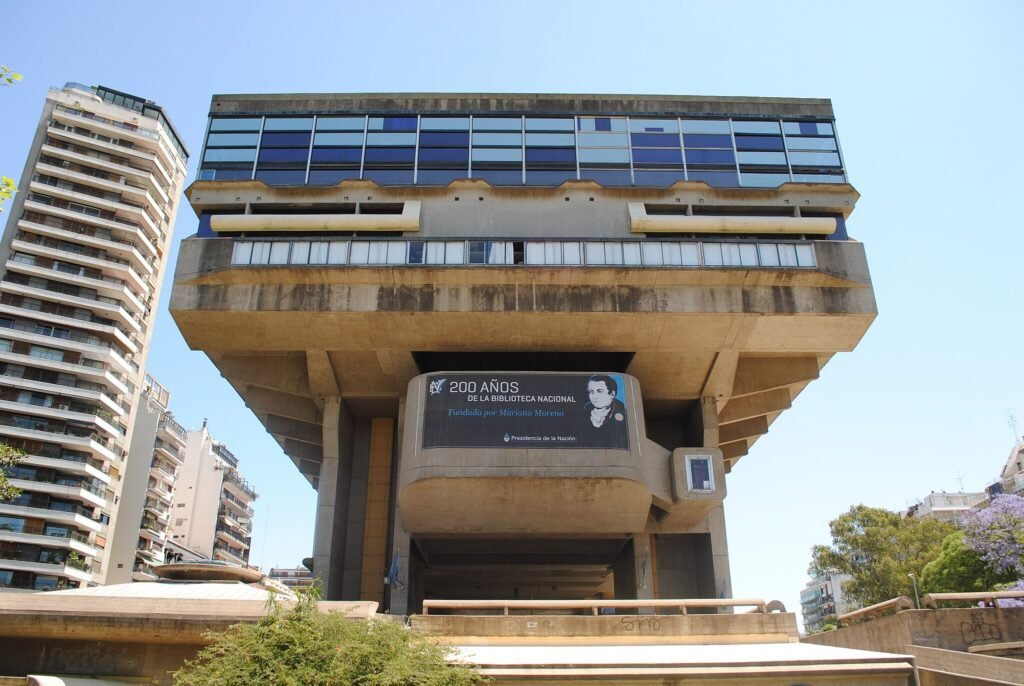A imagem mostra a fachada imponente da Biblioteca Nacional da Argentina, localizada em Buenos Aires. O edifício é um exemplo marcante da arquitetura brutalista, caracterizada pelo uso de concreto aparente e formas geométricas maciças. A estrutura principal da biblioteca se eleva sobre pilares, criando uma sensação de monumentalidade e imponência. Na parte central da fachada, há uma faixa comemorativa destacando os "200 anos da Biblioteca Nacional", com a imagem de Mariano Moreno, um dos fundadores da instituição. O design do prédio contrasta fortemente com os edifícios residenciais modernos ao redor, destacando a importância cultural do local.