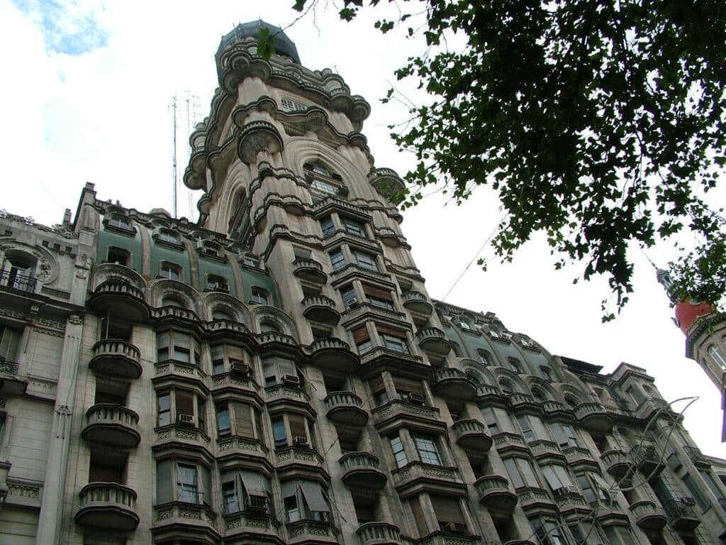 A imagem mostra a fachada do Palacio Barolo, um edifício icônico localizado na Avenida de Mayo, em Buenos Aires. A estrutura destaca-se por seu design arquitetônico imponente, com diversos andares e sacadas decoradas de forma elaborada. O estilo do prédio é eclético, combinando elementos neogóticos e art déco. A parte superior do edifício é dominada por uma cúpula intricada, e há uma árvore cujos galhos aparecem parcialmente na parte superior direita da imagem. O céu está nublado, mas ainda é possível ver detalhes das janelas e varandas do edifício.