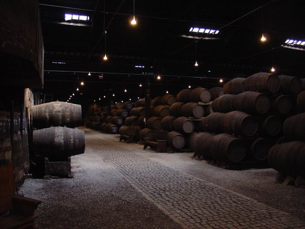 Uma adega de Vinhos do Porto mal iluminada com fileiras de grandes barris de madeira de cada lado de um caminho de paralelepípedos, uma das opções nas Cidades de Portugal