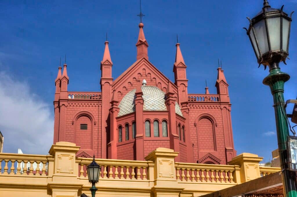 A imagem mostra a fachada do Centro Cultural Recoleta, um edifício icônico localizado no bairro da Recoleta, em Buenos Aires. A construção possui um estilo arquitetônico neogótico, caracterizado por suas torres pontiagudas e detalhes ornamentados. A cor predominante do edifício é um tom avermelhado, com uma cúpula metálica prateada no centro. À frente do edifício, há um parapeito em tom amarelo claro, complementando o contraste de cores com o azul do céu ao fundo. A lanterna decorativa no lado direito da imagem acrescenta um toque de elegância ao cenário urbano.