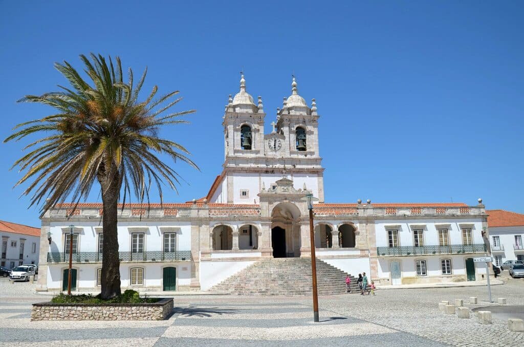 A imagem mostra a Igreja de Nossa Senhora da Nazaré, localizada em Nazaré, Portugal. A igreja possui uma fachada imponente, com duas torres sineiras simétricas, uma em cada lado. As torres são adornadas com detalhes arquitetônicos típicos do estilo barroco, incluindo cúpulas com ornamentos esféricos. A fachada é predominantemente branca com detalhes em pedra, e a entrada principal é acessada por uma escadaria ampla, que leva a um pórtico com colunas e um frontão triangular. Acima do pórtico, há um relógio central, e mais acima, nichos com estátuas religiosas. Em frente à igreja, há uma praça pavimentada com pedras portuguesas, formando padrões geométricos em tons de cinza e branco. Uma palmeira alta está plantada em um canteiro na praça, adicionando um elemento tropical ao cenário. Algumas pessoas podem ser vistas caminhando e apreciando o local.