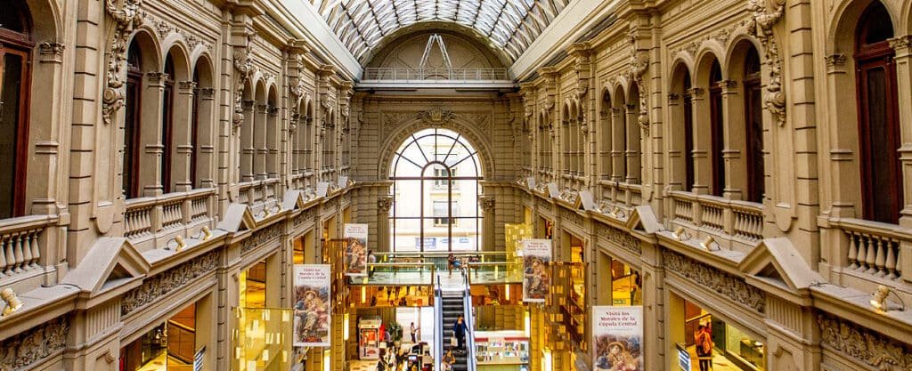 A imagem retrata o interior das Galerías Pacífico, um famoso shopping center localizado em Buenos Aires. O espaço é luxuoso, com arquitetura clássica que remete ao estilo europeu. As paredes são detalhadamente decoradas com ornamentos esculpidos, janelas arqueadas e colunas que dão um ar majestoso ao ambiente. No centro, há um corredor amplo com lojas em ambos os lados e uma escada rolante que leva ao andar superior. O teto de vidro em estilo abobadado permite a entrada de luz natural, iluminando o ambiente de forma suave. Ao fundo, uma grande janela arqueada adiciona profundidade ao espaço, reforçando a sensação de grandiosidade do lugar. Banners e cartazes estão pendurados ao longo do corredor, promovendo eventos ou exposições que ocorrem nas galerias.