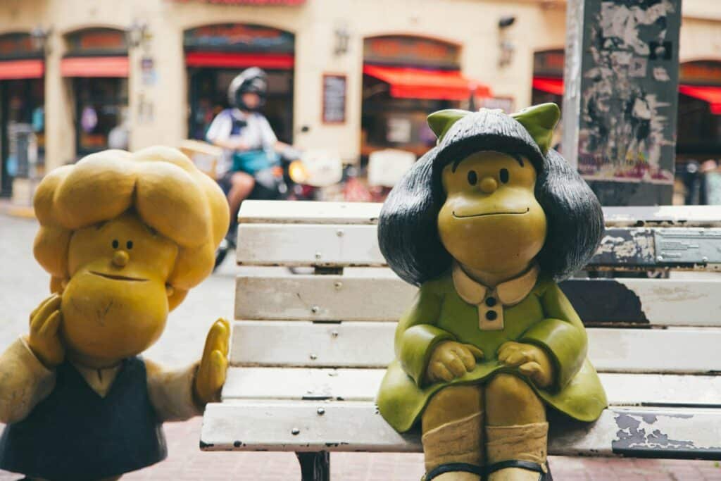 A imagem mostra duas estátuas de personagens da famosa tirinha "Mafalda", criada pelo cartunista argentino Quino. À direita, está Mafalda, uma menina com cabelos pretos e volumosos, vestindo um vestido verde, sentada em um banco branco. Ela tem uma expressão pensativa e está com as mãos apoiadas no colo. À esquerda, vemos Manolito, outro personagem da tirinha, com cabelo loiro e volumoso, usando uma camisa branca e um colete azul. Ele está posicionado ao lado do banco, com uma expressão amigável e um braço levantado, como se estivesse acenando ou cumprimentando alguém. Ao fundo, há uma cena de rua com uma pessoa em uma motocicleta, fachadas de prédios e toldos vermelhos, desfocados, o que dá destaque às estátuas em primeiro plano.
