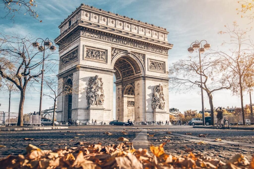 Uma vista de ângulo baixo do Arco do Triunfo em Paris, cercado por árvores e folhas caídas em um dia ensolarado.