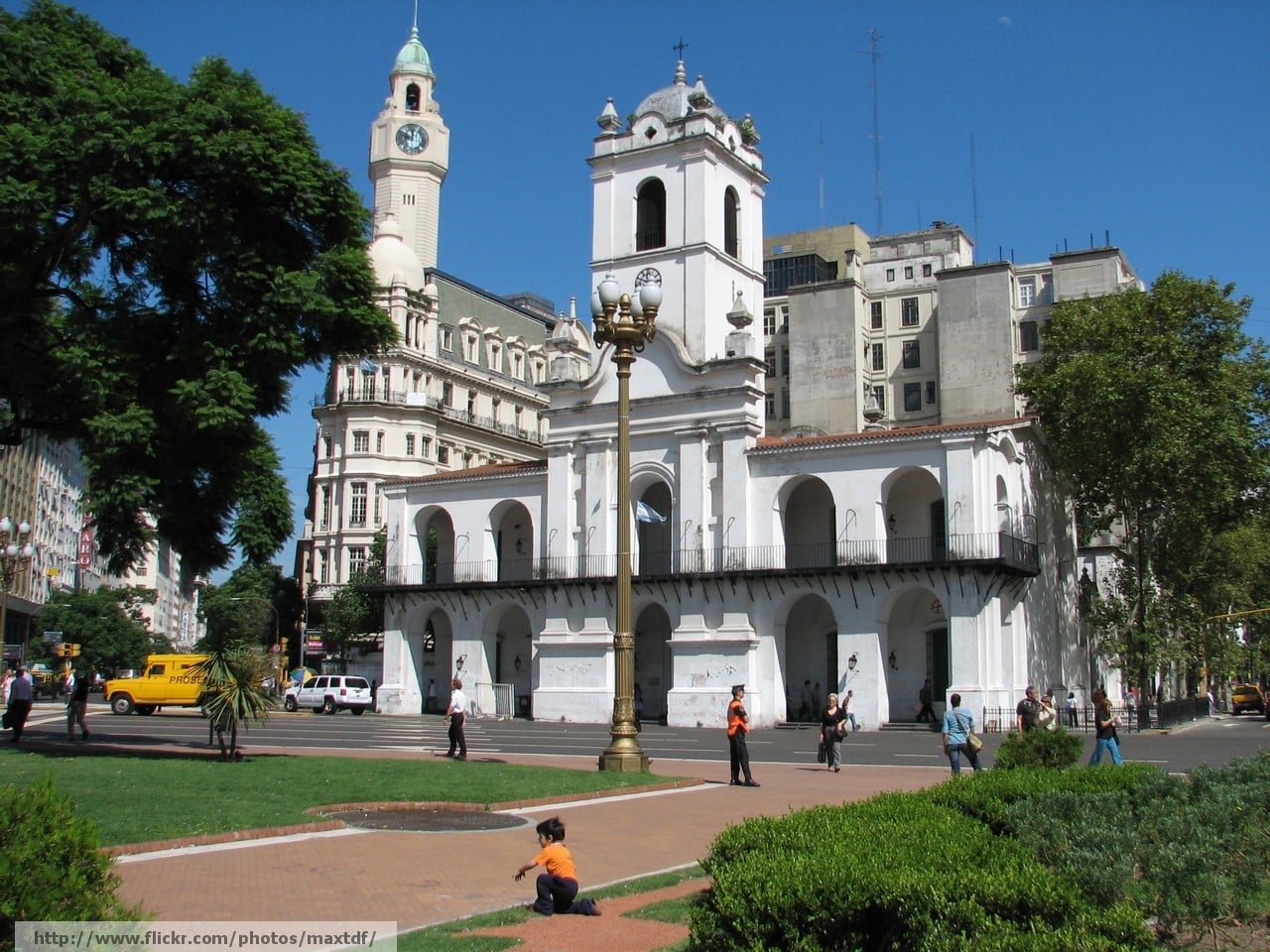 A imagem mostra o Cabildo de Buenos Aires, localizado na Praça de Maio, sob um céu azul claro. O edifício é branco, com uma fachada histórica que exibe uma série de arcos no térreo e no andar superior, além de uma torre com um relógio. À direita, algumas árvores proporcionam sombra ao local, enquanto à esquerda, um edifício de arquitetura clássica com uma torre também é visível. Na praça em frente ao Cabildo, há algumas pessoas caminhando e apreciando o local, enquanto um gramado verde ocupa o primeiro plano da imagem. A cena captura a tranquilidade e a beleza histórica dessa área emblemática de Buenos Aires.