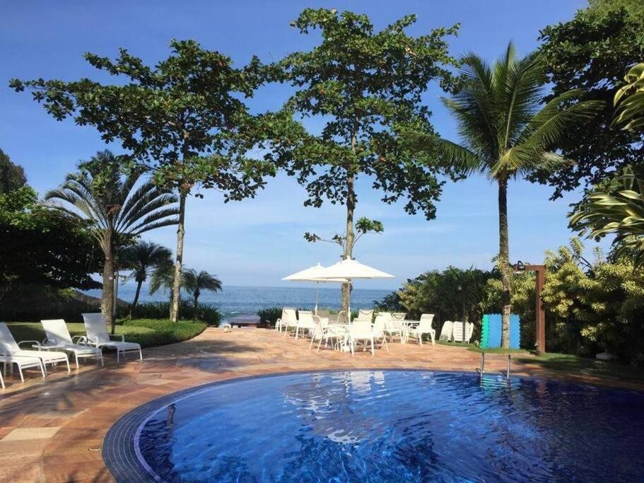 Piscina da Casa Deliciosa em Condo c/ Piscina Frente ao Mar cercada por árvores, espreguiçadeiras e mesinhas com cadeiras e guarda-sóis. Ao fundo é possível ver o mar.