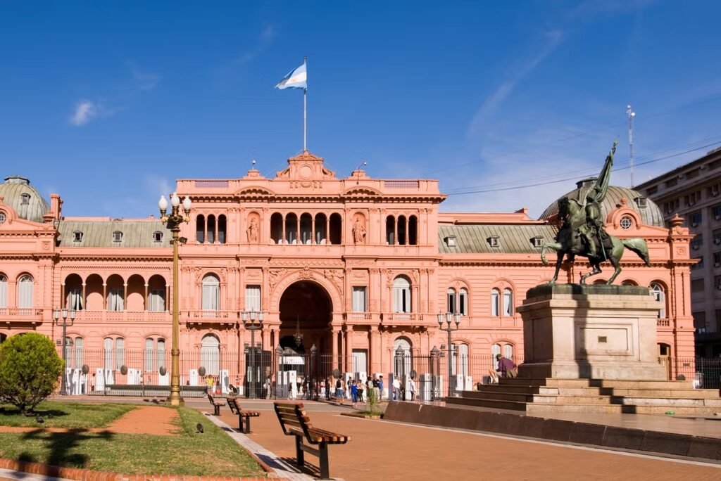A imagem retrata a Casa Rosada, o icônico palácio presidencial da Argentina, localizado em Buenos Aires. O edifício é caracterizado por sua cor rosada distintiva e arquitetura neoclássica, com detalhes ornamentados e arcadas. No centro, há uma grande entrada em arco, e a fachada é adornada com janelas altas e varandas. Sobre o edifício, uma bandeira argentina tremula no topo. À direita da imagem, uma estátua equestre de um herói nacional, provavelmente o General Manuel Belgrano, destaca-se em um pedestal. A imagem foi capturada sob um céu azul claro, em um dia ensolarado, com alguns bancos e áreas verdes no primeiro plano, criando uma cena serena e representativa deste marco histórico argentino.