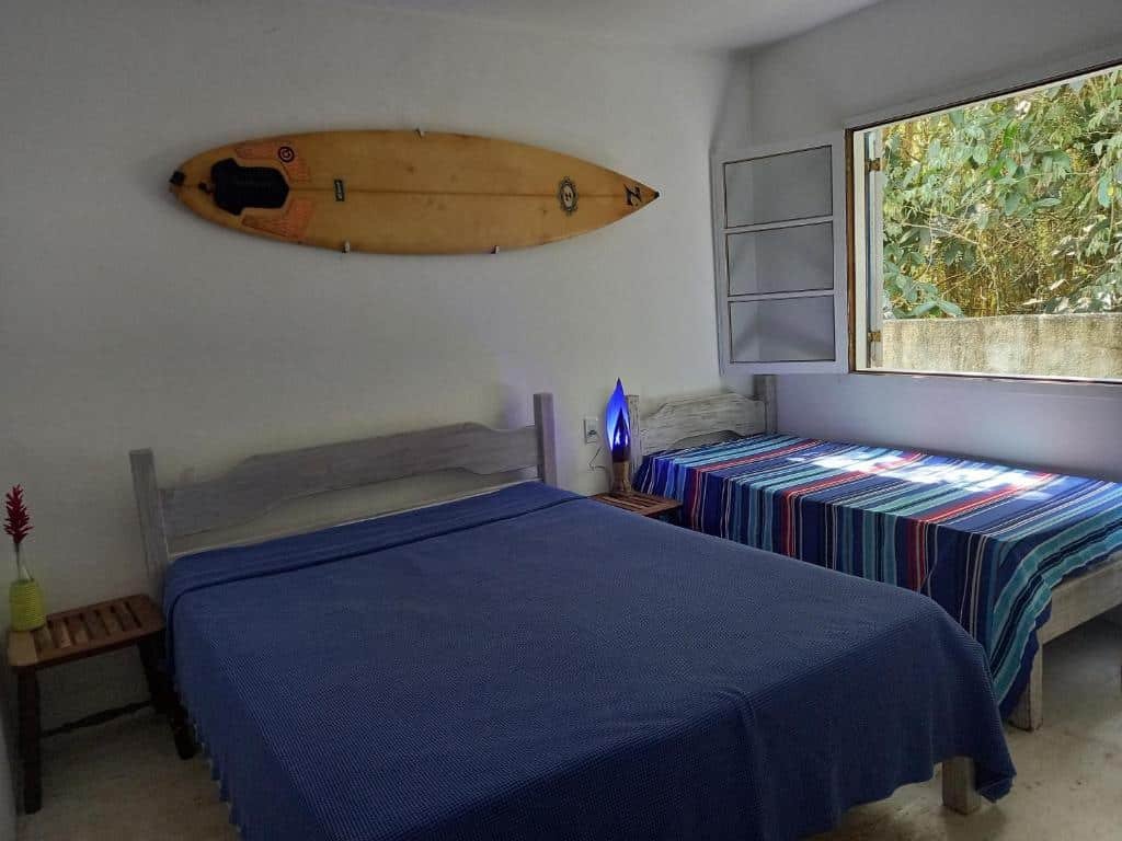 Quarto da Casa sossegada ao lado da natureza e do mar, um dos airbnb em Camburizinho. Uma cama de casal está no centro do quarto com mesinhas de cabeceira dos dois lados, e na lateral direita há uma cama de solteiro encostada na parede da janela. Na parede acima das camas há uma prancha pendurada.