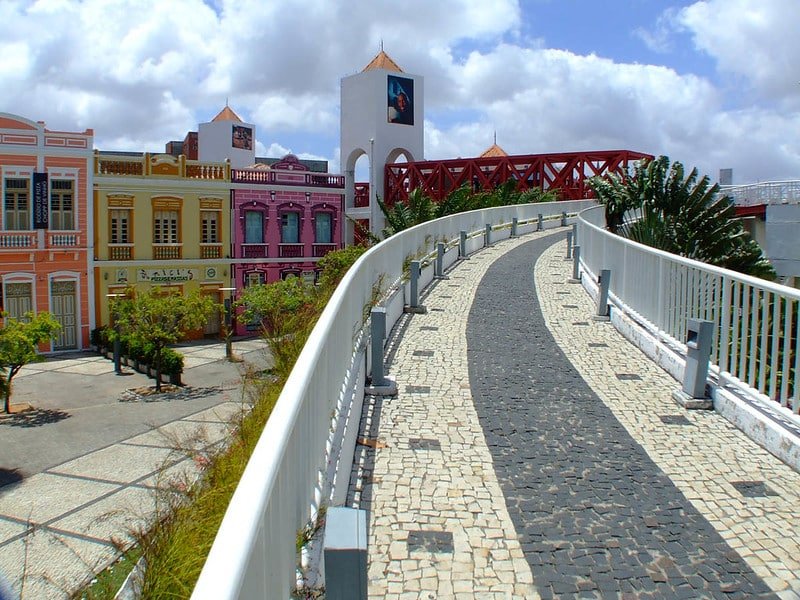 A imagem mostra uma passarela com um corrimão branco que leva até a passarela do Centro Dragão do Mar de Arte e Cultura, um dos pontos turísticos de Fortaleza. A ponte é vermelha feita de concreto e possui arcos de metal. Na direita, no fundo a foto, vê-se prédios coloridos.