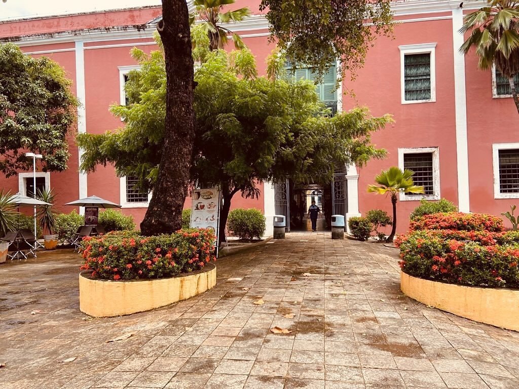 A imagem mostra um pátio com jardim e um prédio é de três andares, com janelas retangulares. Há uma placa na frente do prédio com a inscrição "Centro de Turismo do Ceará (Emcetur)", um dos pontos turísticos em Fortaleza.