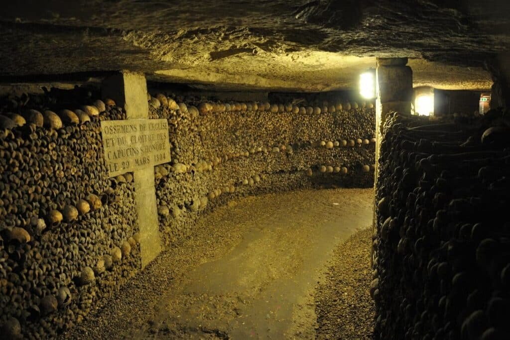 Catacumbas subterrâneas mal iluminadas com paredes de crânios e ossos humanos empilhados, com uma placa e pilares de pedra. Esse é um dos pontos turísticos de Paris.