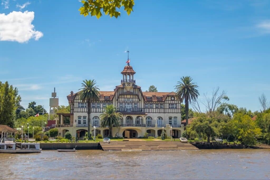 A imagem retrata o Clube de Regatas La Marina, localizado às margens do rio Tigre, na região do Delta do Paraná, em Buenos Aires. A construção é imponente e de estilo arquitetônico clássico, com uma combinação de elementos coloniais e europeus. A fachada do edifício é simétrica, destacando-se por seus detalhes em madeira escura que contrastam com as paredes de cor clara.
O edifício possui três andares, com varandas no segundo e terceiro pisos, e um telhado de estilo colonial com várias águas, adornado com uma pequena torre central. À frente do clube, palmeiras e outras árvores proporcionam um ambiente tropical, complementando a atmosfera serena à beira do rio.