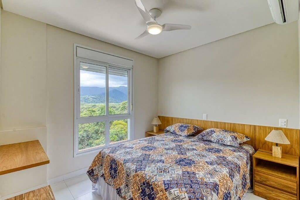 Quarto do D16 - Conforto junto a natureza, um dos airbnb em Camburizinho. Uma cama de casal tem mesinhas de cabeceira e abajures dos dois lados, assim como um ventilador de teto logo acima. Na parede esquerda fica uma janela com vista, e na parede direita há um aparelho de ar-condicionado.