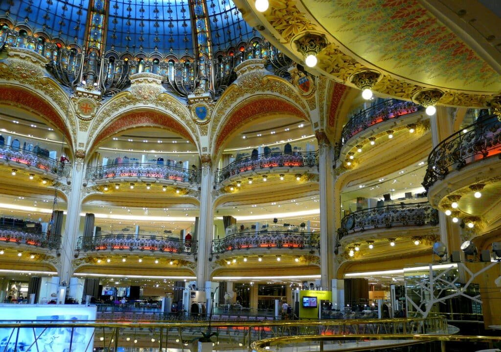 Interior de um shopping center de vários andares com varandas ornamentadas, um grande teto de vidro abobadado e iluminação decorativa.