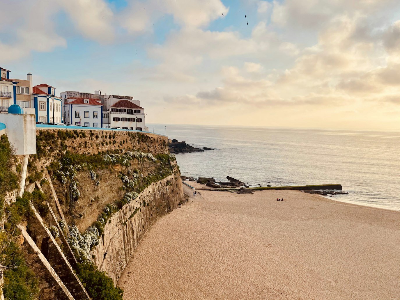 Cena costeira em Ericeira com casas no topo de um penhasco rochoso, com vista para uma praia de areia e mar calmo sob um céu parcialmente nublado.