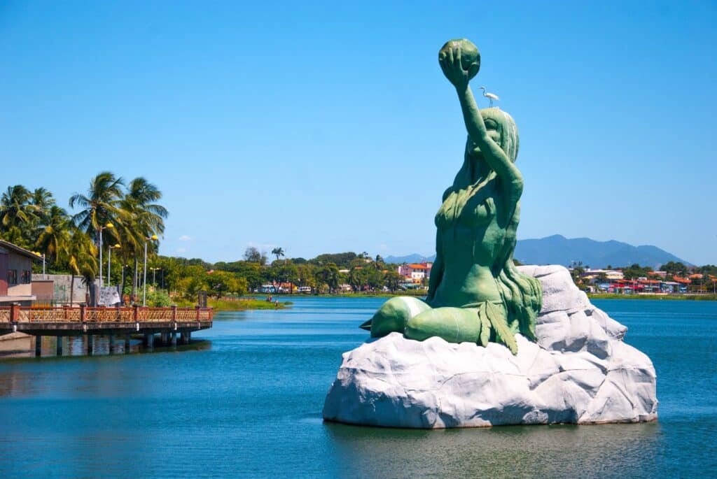 A imagem foi tirada em Fortaleza, e mostra uma estátua de uma sereia sentada em cima de uma rocha em meio a um lago. A sereia é uma figura feminina com cauda de peixe. Ela está sentada em uma rocha com as pernas cruzadas e os braços apoiados nos joelhos. A sereia está olhando para frente com uma expressão serena no rosto.

Ao redor da sereia, há água calma e clara. No fundo, há árvores e outras plantas aquáticas. O céu está azul e limpo, com algumas nuvens brancas no horizonte. É um dos pontos turísticos em Fortaleza.