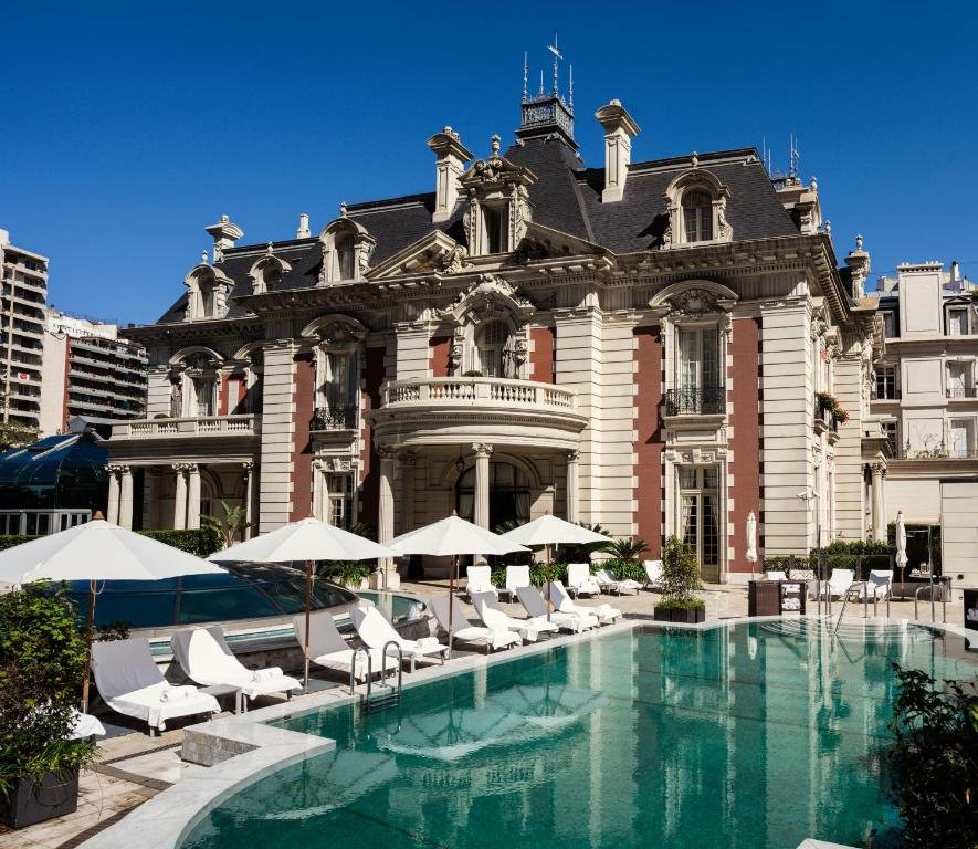 A imagem mostra um dos hotéis para família em Buenos Aires, o Four Seasons Hotel Buenos Aires. A fachada combina arquitetura clássica europeia com elementos modernos. Em primeiro plano, há uma piscina cercada por espreguiçadeiras brancas e guarda-sóis, proporcionando um ambiente de relaxamento ao ar livre. O prédio principal do hotel é uma mansão histórica de estilo francês, com detalhes arquitetônicos sofisticados, como colunas e molduras ornamentadas nas janelas. O céu está claro e azul, destacando ainda mais a imponência da construção. Ao fundo, é possível ver outros edifícios que contrastam com o estilo clássico da mansão.