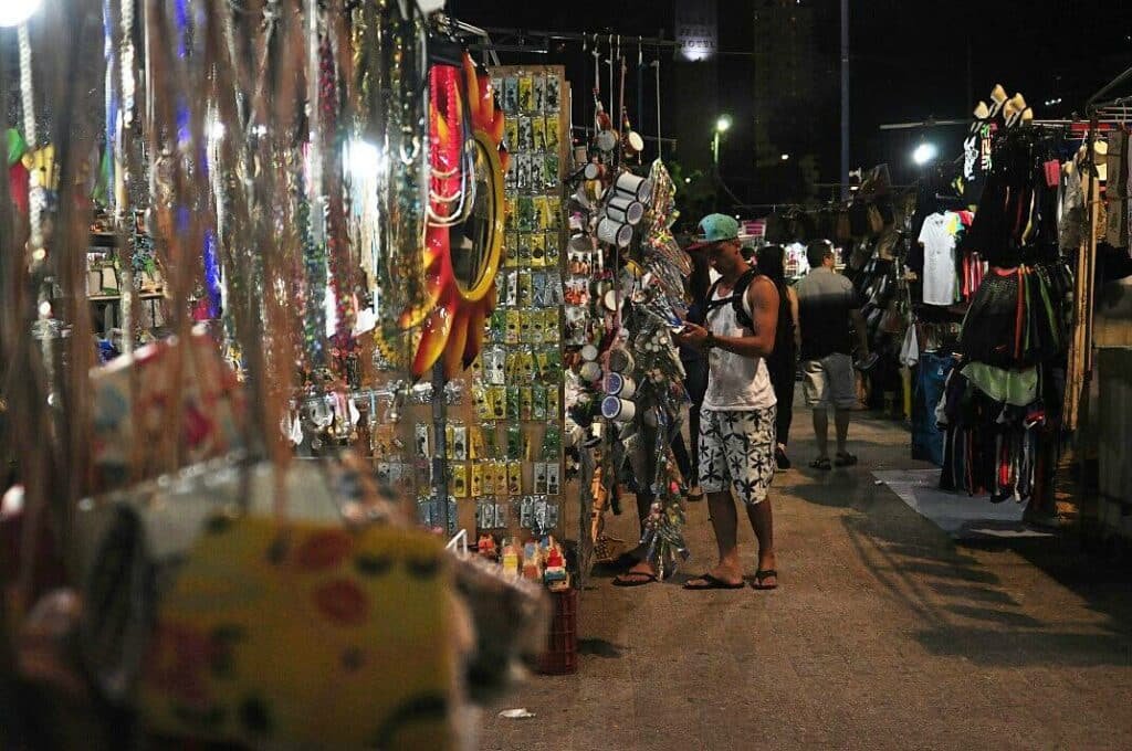 Foto tirada a noite na Feirinha de Artesanato Beira-Mar, em Fortaleza. Há barraquinhas com peças artesanais. Um homem está olhando os itens. Há outras pessoas no fundo, caminhando entre as barracas.