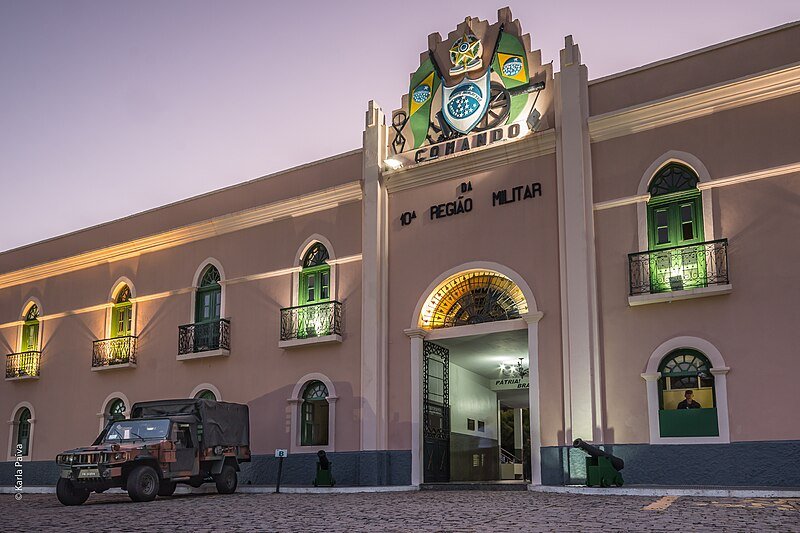 A imagem mostra um caminhão militar estacionado em frente a um prédio. O caminhão é verde e tem um logotipo militar na porta. O prédio é branco e tem três andares. Há uma placa na frente do prédio com a inscrição "COMANDO DA 10ª REGIÃO MILITAR". É o prédio da Fortaleza Nossa Senhora de Assunção, um dos pontos turísticos em Fortaleza.