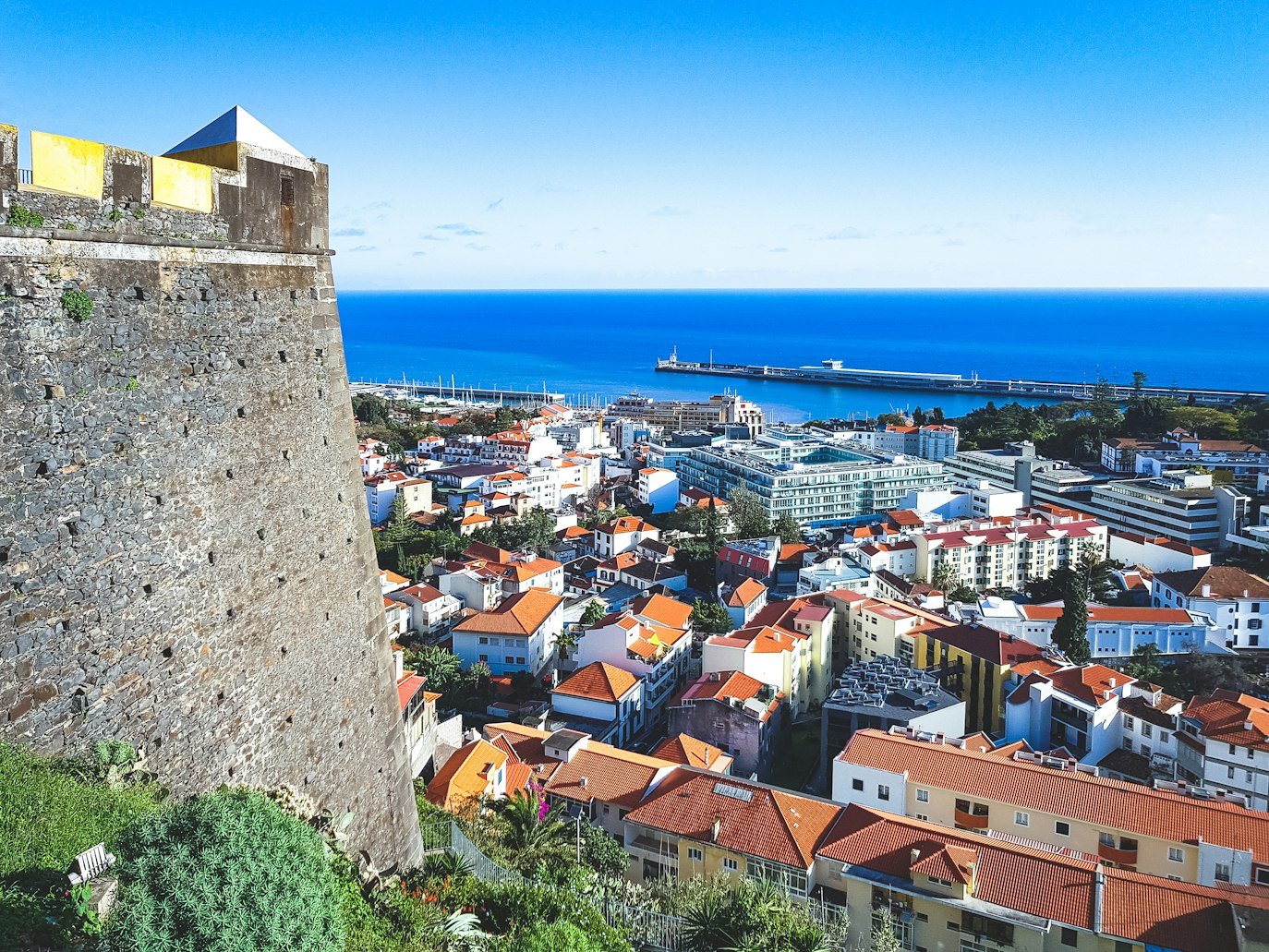 Vista elevada de uma paisagem urbana costeira com edifícios coloridos, um grande muro de pedra e um porto visível contra um céu azul claro.
