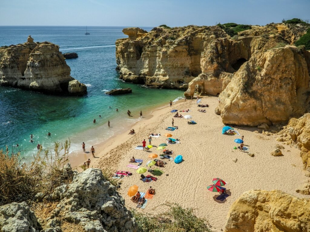 Vista aérea de uma praia no Algarve, em Portugal. O local é cercado por grandes rochas, e há banhistas relaxando na areia. Na lateral esquerda fica um pedaço do mar cristalino.