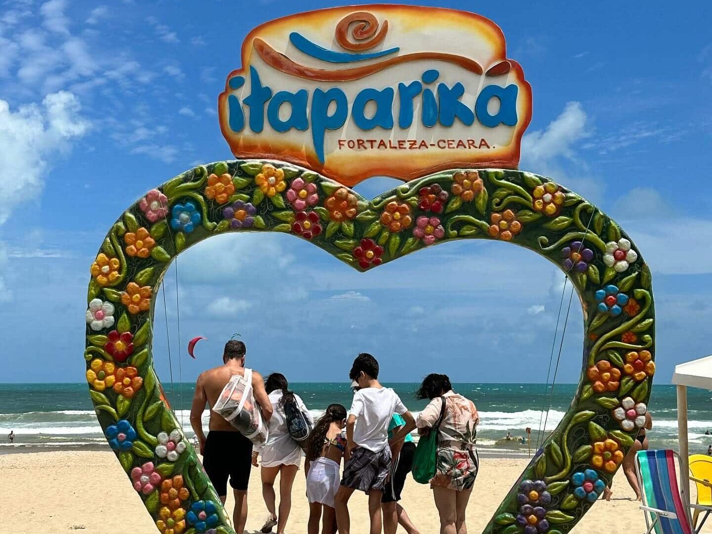 A foto foi tirada de dia e mostra um arco em formato de coração com uma placa em cima escrito "Itapariká, Fortaleza - Ceará". Debaixo do arco há uma família de costas para a foto. No fundo está o mar.