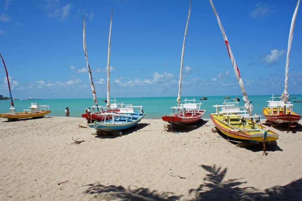 Jangadas de diferentes 
 cores estacionadas na areia branca com um mar cristalino ao fundo. Há sombra de coqueiros à frente e a imagem foi tirada em um dia ensolarado. Foto para ilustrar o post de o que fazer em Maceió