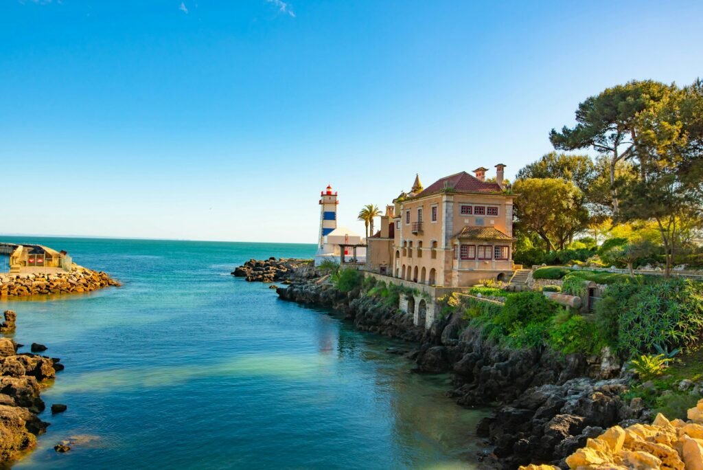 Mar próximo ao Farol de Santa Marta em Cascais, Portugal. Um sobrado fica ao lado do farol em cima de um terreno de pedregulhos. Há várias árvores ao redor.