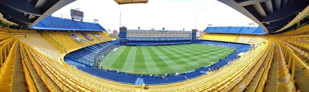 A imagem mostra o estádio La Bombonera, casa do clube de futebol Boca Juniors, localizado em Buenos Aires, Argentina. O estádio é conhecido por suas arquibancadas íngremes e sua capacidade de criar uma atmosfera vibrante durante os jogos. As arquibancadas são predominantemente amarelas e azuis, as cores tradicionais do clube. O campo de futebol está bem cuidado, com a grama cortada em faixas. A estrutura do estádio, com suas arquibancadas quase verticais, é projetada para maximizar a proximidade dos torcedores ao campo, o que contribui para a famosa intimidade e intensidade do local durante as partidas.
