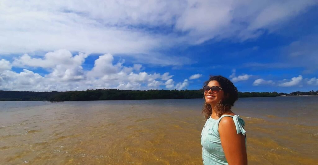 Mulher olhando para o lado, com o cabelo cacheado solo, com a lagoa do Roteiro, na praia do Gunga, de fundo e árvores ao fundo. Representa o que fazer em Maceió