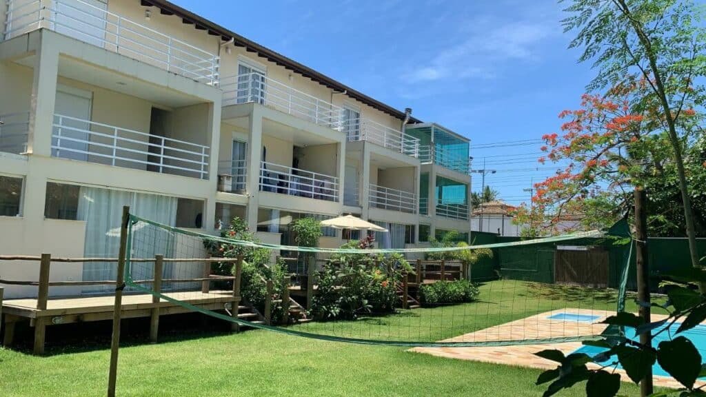 Vista da fachada da Linda Casa em Camburizinho 100 m da praia, que tem sacadas com grades brancas. Logo em frente há um jardim com plantas e piscina ao lado de uma rede de vôlei.