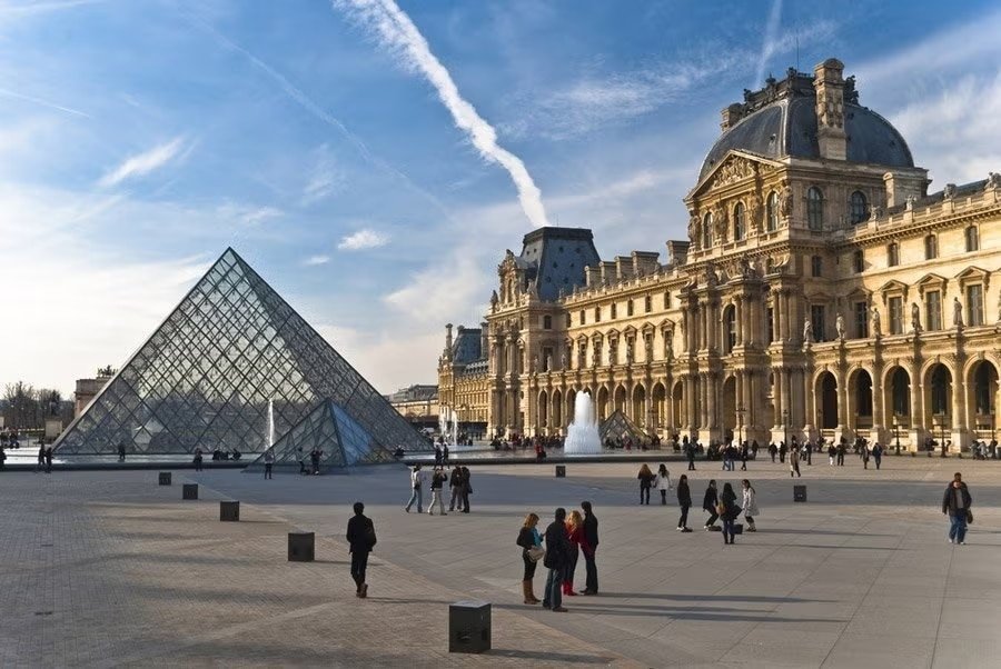 Pessoas caminhando perto da Pirâmide do Louvre com o histórico Palácio do Louvre ao fundo e um céu azul claro acima.