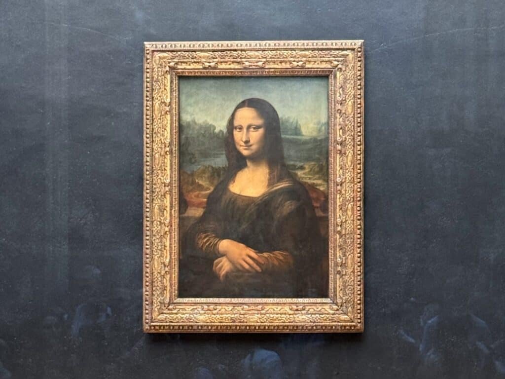 Uma pintura emoldurada da Mona Lisa com um fundo escuro. A obra de arte apresenta uma mulher com um sorriso sutil e mãos postas.