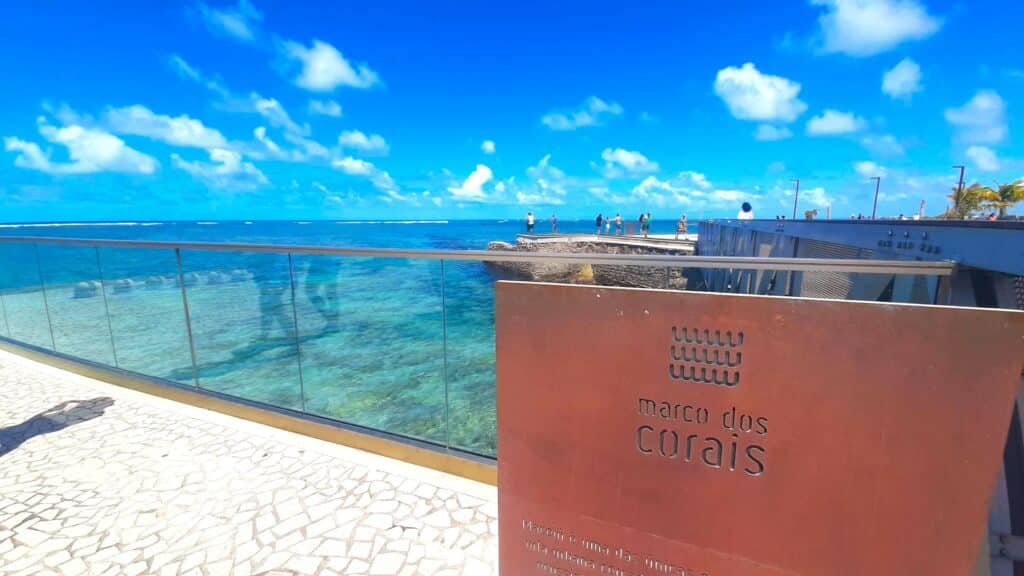 Placa escrito "Marco dos Corais" com uma parede baixa de vidro e o mar de fundo. O local é um dos pontos turísticos em Maceió