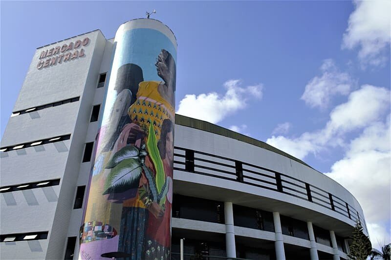 A imagem mostra um mural pintado na parede lateral de um prédio. O mural é colorido e retrata uma cena urbana com prédios altos, ruas movimentadas e pessoas. No canto superior esquerdo do mural, há um letreiro com o nome "Mercado Central".