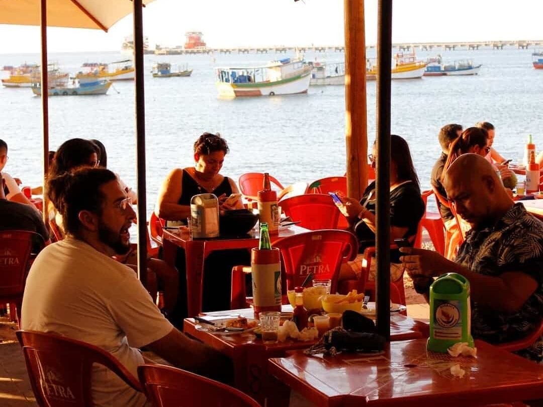 Pessoas sentadas em mesas e cadeiras de bar no Mercado dos Peixes, em Fortaleza. Estão debaixo de guarda-sóis enquanto conversa, comem petiscos e bebem cerveja. No fundo está o mar com algumas embarcações.