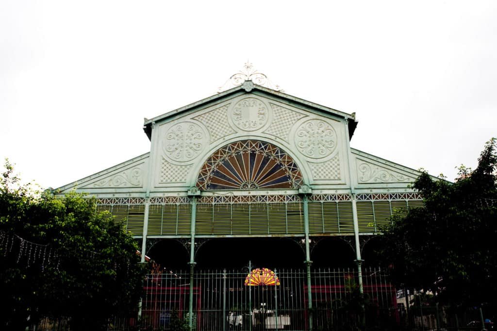 Foto tirada em um dia nublado, de frente para o Mercado dos Pinhões em Fortaleza. O prédio possui portões de ferro e um vitral colorido perto do telhado. Há árvores em seu entorno. É um dos pontos turísticos em Fortaleza.