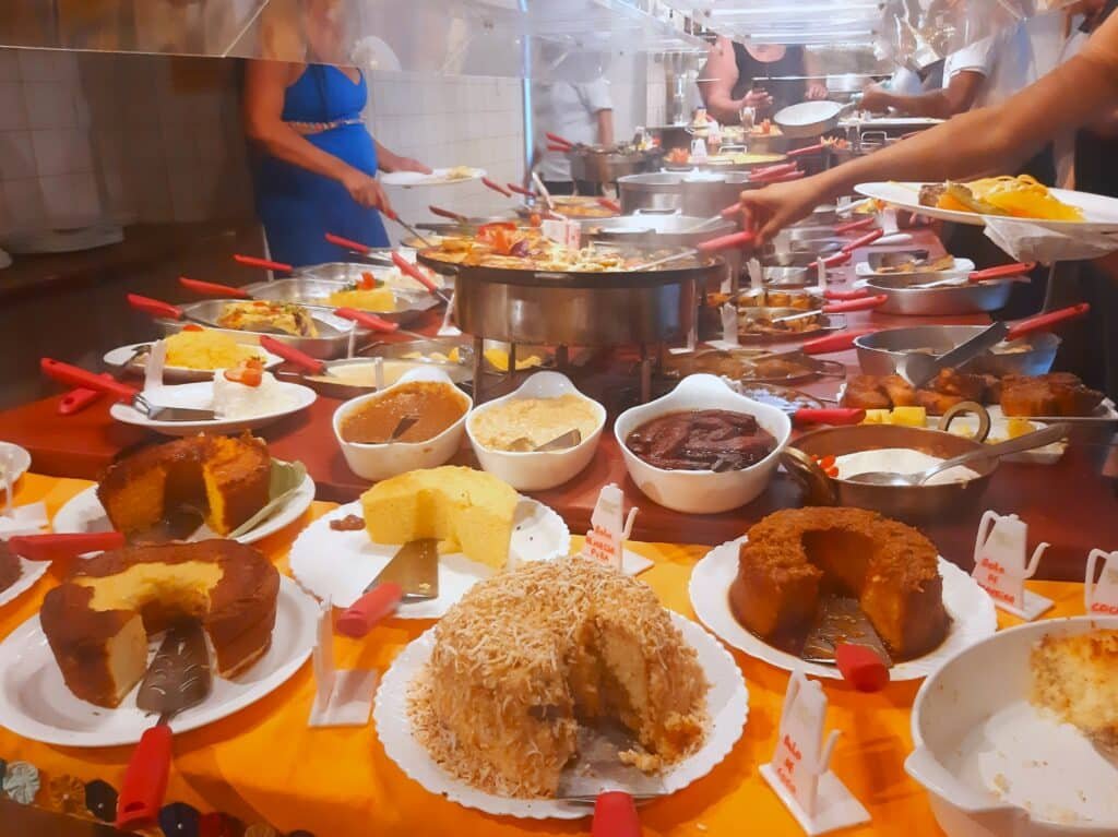 Mesa self service com diferentes tipos de bolos e doces e panelas com receitas salgadas ao fundo. Há algumas pessoas se servindo com pratos e colher na mão