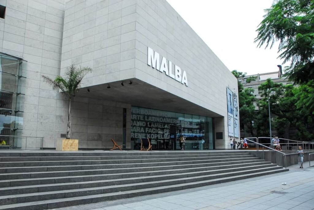 A imagem mostra a fachada do Museu de Arte Latino-Americana de Buenos Aires (MALBA). O edifício possui uma arquitetura moderna, com linhas retas e uma estrutura predominantemente de concreto e vidro. A entrada principal é acessada por uma ampla escadaria, e a sigla "MALBA" está em destaque na parte superior da fachada. O design é minimalista e elegante, refletindo a natureza contemporânea do museu, que é um importante centro cultural dedicado à promoção e preservação da arte latino-americana. A área ao redor é arborizada, acrescentando um toque de verde ao ambiente urbano.