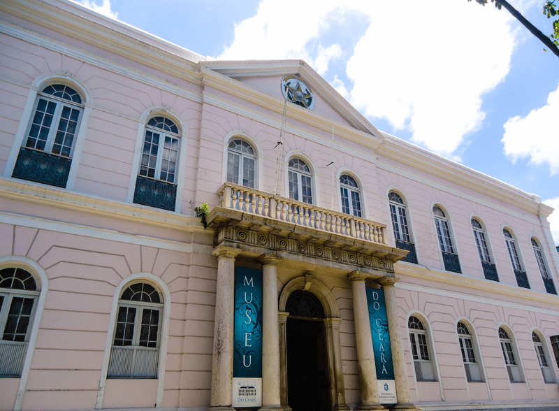 A foto mostra a fachada do Museu do Ceará, um dos pontos turísticos em Fortaleza. O prédio possui arquitetura antiga conservada, com dois andares. Há várias janelas e uma porta central alta.