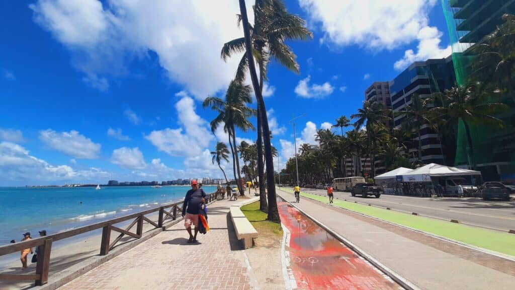 Orla de Jatiúca, uma das opções de o que fazer em Maceió. Do lado esquerdo tem um mar azulado e, no esquerdo, um calçadão com um senhor andando, alguns coqueiros e uma avenida com carros e prédios ao fundo