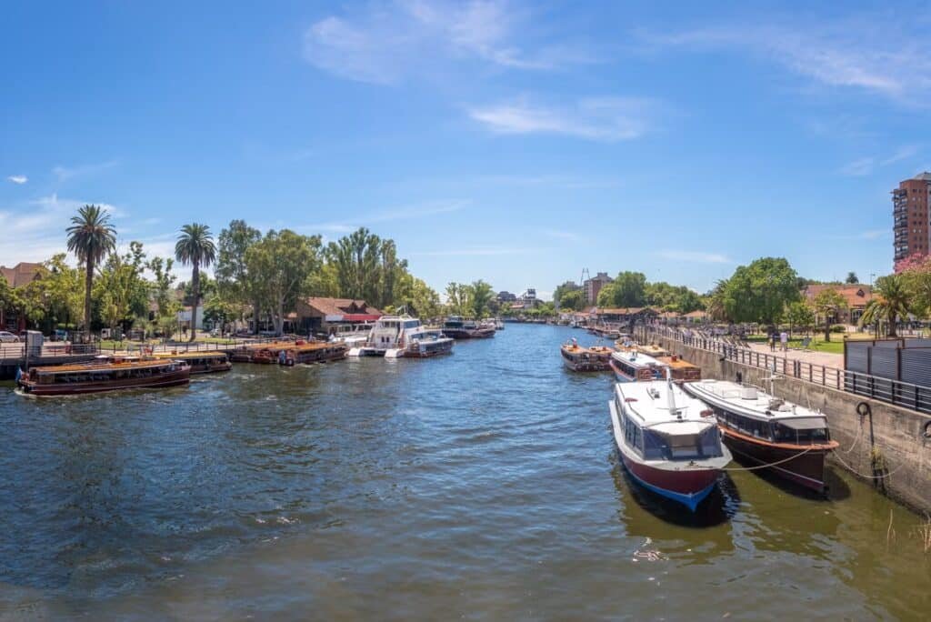 A imagem mostra um cenário do Rio da Prata, em Buenos Aires, com diversos barcos ancorados ao longo da margem. A água do rio reflete o céu azul claro, sem nuvens significativas. Nas margens, há áreas arborizadas com palmeiras e outros tipos de árvores, e uma calçada que segue paralela ao rio, onde pessoas caminham e desfrutam do ambiente. Ao fundo, é possível ver algumas edificações e vegetação densa. O clima ensolarado e as condições do local sugerem um ambiente agradável para passeios de barco e atividades ao ar livre.
