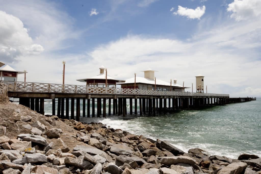 A imagem mostra o píer da Ponte dos Ingleses, um dos pontos turísticos em Fortaleza. É extenso e se estende para o oceano. O píer é feito de madeira e possui um telhado branco. Ele é cercado por rochas e ondas quebrando na costa.