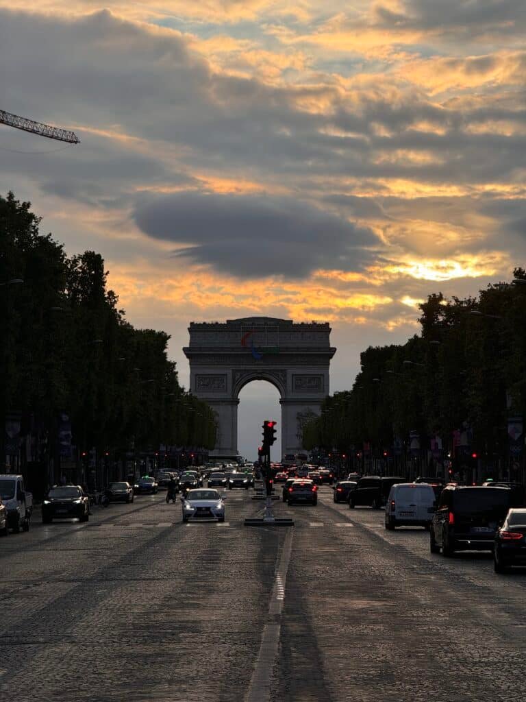 Uma vista do pôr do sol no Arco do Triunfo com carros e árvores ao longo da rua em uma rua movimentada de Paris. Esse é um dos pontos turísticos de Paris.