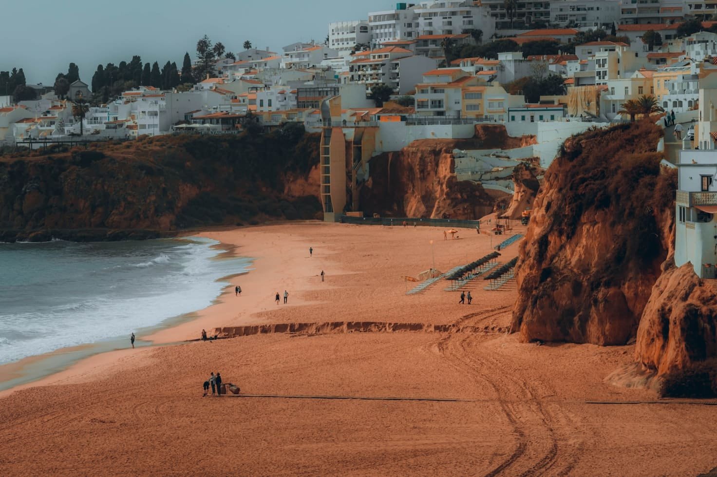 Praia de areia em Portimão com poucas pessoas, cercada por penhascos e prédios brancos. O oceano fica à esquerda e degraus levam até a praia.
