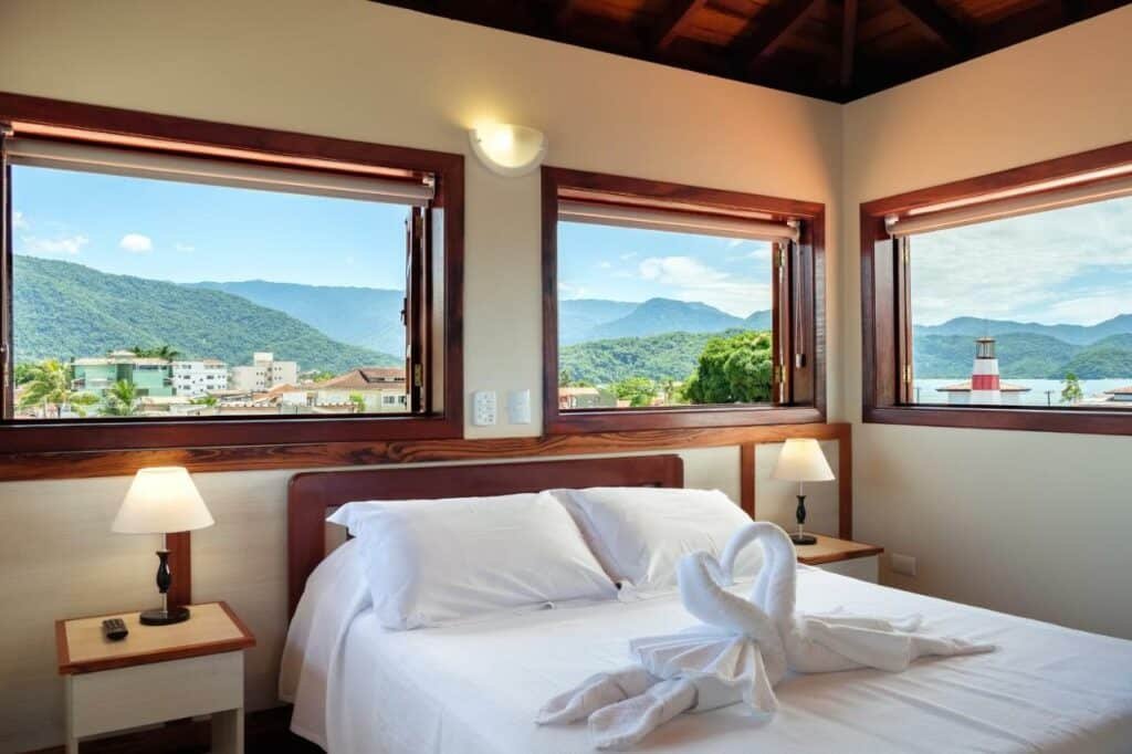 Vista das montanhas que cercam a cidade a partir das janelas da Pousada Don Diego, com uma cama de casal e toalhas em formato de cisne sobre ela