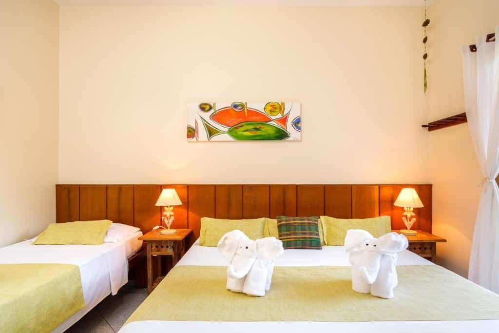 Quarto com uma cama de solteiro e uma cama de casal com mesas de cabeceira e abajures em ambos os lados. Há toalhas de banho em formato de elefante sobre a cama maior