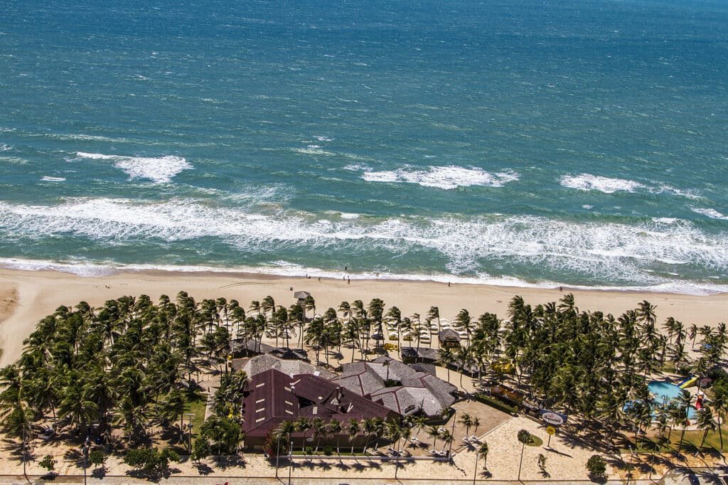 Vista aérea da Praia do Futuro, uma das melhores Praias de Fortaleza. Vê-se a areia branca, coqueiros, uma construção com vários quiosques e o mar.