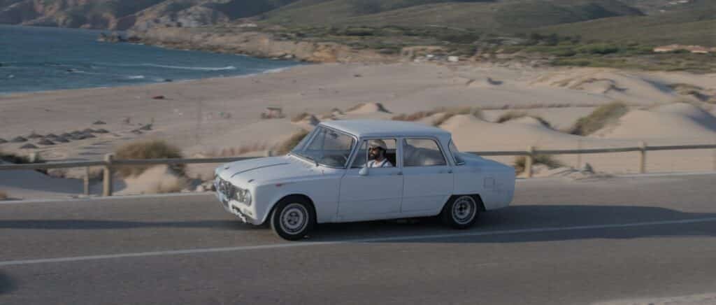Um carro branco antigo passa por uma estrada costeira com uma praia de areia e penhascos rochosos ao fundo.
